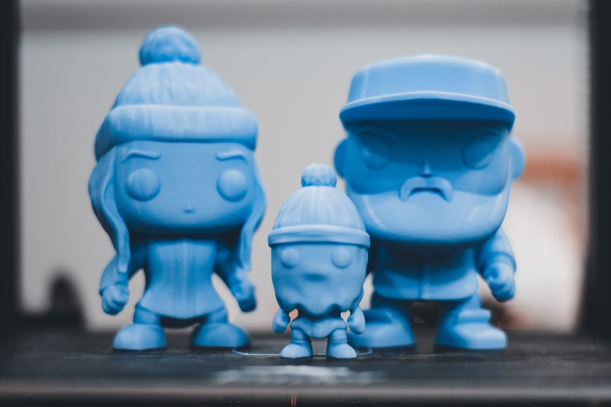 Tres figuras de personajes impresas en 3D en filamento celeste, recién salidas de la impresora.