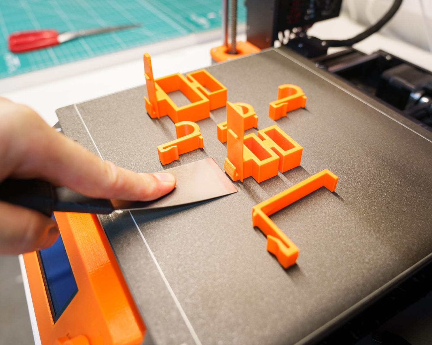 Una mano retira con una espátula varias piezas naranjas recién impresas de la cama de la impresora 3D.