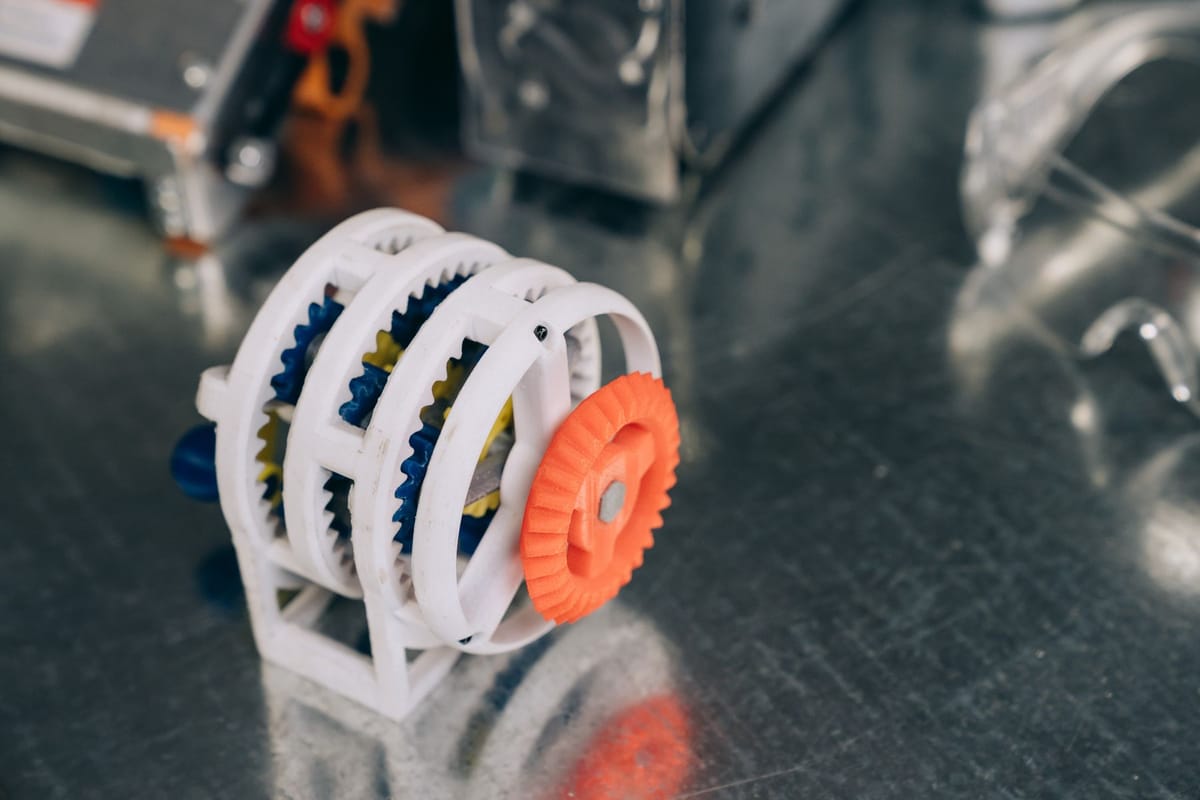Prototipo de un mecanismo de engranajes impreso en 3D en blanco, naranja y azul sobre la mesa de trabajo.
